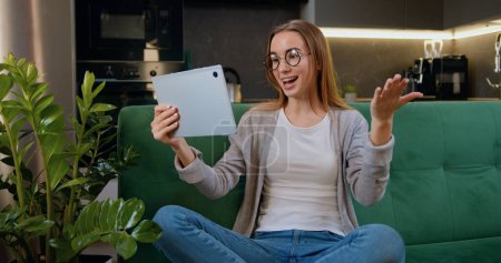 Attraktive junge Frau, die auf dem Sofa sitzt und im Hintergrund in der Küche einen Videochat auf dem Tablet-Gerät führt. Menschen, Mädchen, Online-Kommunikationskonzept