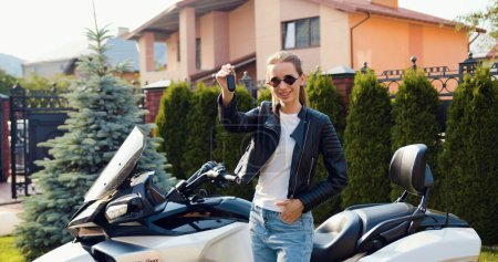 Téléchargez les photos : Heureux attaractive femme motard dans les lunettes de soleil montrant les clés debout sur sa nouvelle moto à l'extérieur dans la rue. - en image libre de droit