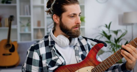 Photo for Close up of caucasian young bearded man with headphones having online electric guitar class at home with laptop in front on table - Royalty Free Image