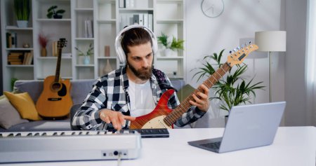 Photo for Musician man in headphones created electronic music on synthesizer piano and e guitar sitting at table at home. Male creating music using equalizer program, recording song in home record studio - Royalty Free Image