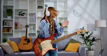 Photo for Energetic funny girl with long hair playing electric guitar, sing song and dancing at living room at home. Attractive girl rocker of 11-12 years old playing rock music on electric guitar. - Royalty Free Image