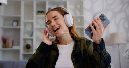 Téléchargez les photos : Gros plan d'une jolie jeune femme riante écoutant de la musique avec smartphone et écouteurs sans fil sur la pièce lumineuse, à l'intérieur - en image libre de droit