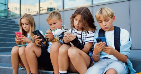 Téléchargez les photos : Écoliers tenanciers de deux filles et garçons caucasiens tenant un téléphone portable et naviguant assis sur un escalier pendant les vacances scolaires, à l'extérieur - en image libre de droit