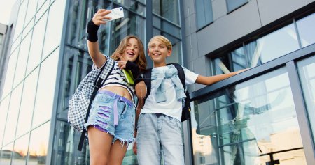 Cute funny small teens boy and girl standing near school building with books and backpacks while doing mocking faces to smartphone camera while taking selfie photos. School concept.