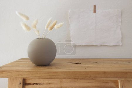 Leere Papierbögen und Poster-Attrappen, die auf eine weiße Wand geklebt sind. Holzbank, Tisch vorhanden. Moderne weiße Keramikvase mit trockenem Lagurus ovatus Gras und Tasse Kaffee. Skandinavisches Landesinnere. Selektiver Fokus.