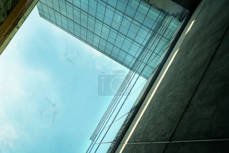 Téléchargez les photos : Vue de bas en haut de l'immeuble de bureaux en verre durable moderne et avion volant dans le ciel bleu. Vue extérieure de l'architecture des bâtiments en verre du siège social de l'entreprise. Bâtiment économe en énergie. Vitre en verre - en image libre de droit