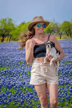 Foto de Una hermosa modelo morena disfruta de un campo de flores Bluebonnet con su perro en un día de primavera - Imagen libre de derechos