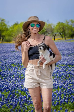 Foto de Una hermosa modelo morena disfruta de un campo de flores Bluebonnet con su perro en un día de primavera - Imagen libre de derechos