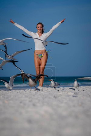 A beautiful blonde bikini model enjoys the weather outdoors on the beach while chasing seagulls in the foreground