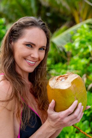 Sun-kissed brunette, adorned in a bikini, revels in Caribbean bliss on the sandy shores, combining beauty with adventure while hunting for coconuts