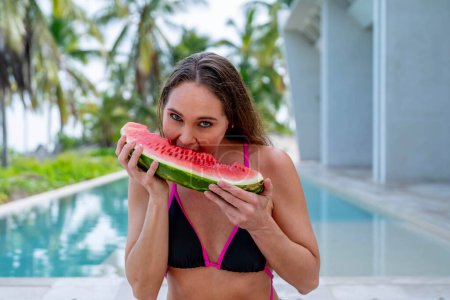 Radiant by the pool, a young beauty basks in vacation bliss. Her smile glistens as she savoringly devours watermelon, epitomizing carefree summer allure.