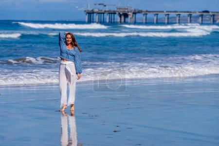 Breathtaking woman basks in beach bliss, near a pier, under clear blue skies, embracing the serenity of a perfect day by the sea