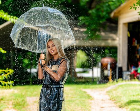 Photo for A radiant blonde model gracefully poses in the rain, donning a raincoat and holding an umbrella, her smile adding charm to the spring scene. - Royalty Free Image