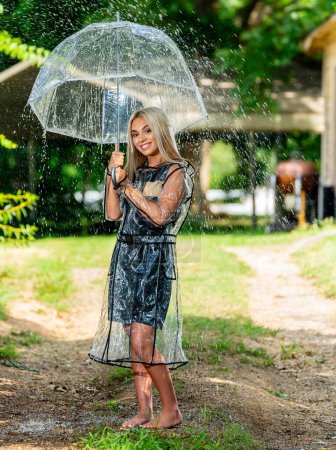 A radiant blonde model gracefully poses in the rain, donning a raincoat and holding an umbrella, her smile adding charm to the spring scene.
