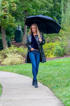 Eine atemberaubende Frau schlendert durch den Park, ihre Eleganz wird durch einen schwarzen Regenschirm akzentuiert, inmitten der drohenden Dunkelheit, bevor der Regen die Erde küsst.