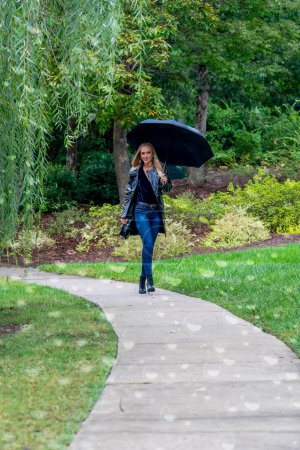 Eine atemberaubende Frau schlendert durch den Park, ihre Eleganz wird durch einen schwarzen Regenschirm akzentuiert, inmitten der drohenden Dunkelheit, bevor der Regen die Erde küsst.