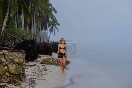 Allein an einem karibischen Strand flaniert eine atemberaubende Frau an der nebelverhangenen Küste und verkörpert Einsamkeit und Schönheit in der ruhigen Umarmung eines Alleinurlaubs