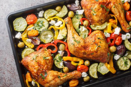 Greek style baked chicken quarters with zucchini, tomatoes, peppers, olives and feta cheese close-up on a baking sheet on the table. Horizontal top view from abov