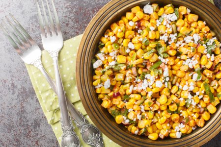 Esquites salad is a grilled Mexican corn with a creamy sauce, seasoned with chile and lime juice and topped with Cotija cheese close-up on the plate on the table. Horizontal top view from abov