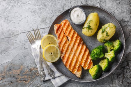 Dietary grilled salmon with boiled potatoes, broccoli, lemon, herbs and cream sauce close-up in a plate on the table. Horizontal top view from abov