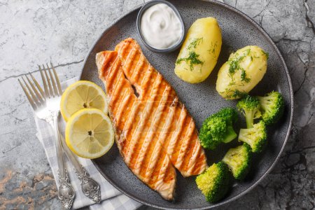 Delicious grilled salmon steak with boiled new potatoes, broccoli, lemon and cream sauce close-up in a plate on the table. Horizontal top view from abov