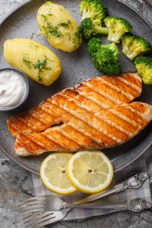 Portion of grilled salmon with boiled potatoes, broccoli, lemon, herbs and cream sauce close-up in a plate on the table. Vertical top view from abov