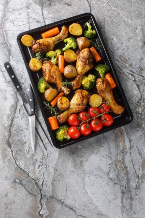 Homemade baked chicken drumsticks with vegetables and rosemary close-up on a baking sheet on the table. Vertical top view from abov