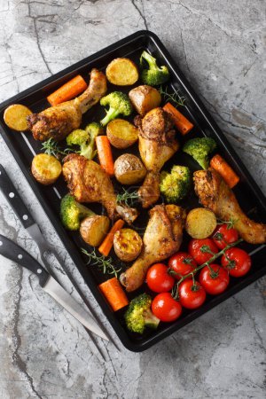 Baked chicken drumstick with broccoli, potatoes, tomatoes, and carrots close-up on a sheet pan on the table. Vertical top view from abov
