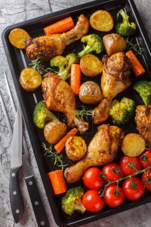 Healthy food baked chicken drumstick with broccoli, potatoes, tomatoes and carrots close-up on a sheet pan on the table. Vertical top view from abov