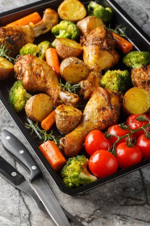 Baked chicken drumstick with broccoli, potatoes, tomatoes, and carrots close-up on a sheet pan on the table. Vertica