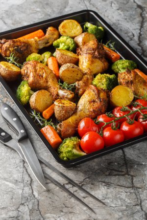 Healthy food baked chicken drumstick with broccoli, potatoes, tomatoes and carrots close-up on a sheet pan on the table. Vertica