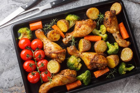 Tasty Baked ruddy chicken legs with potatoes, broccoli, tomato, carrot close-up on a baking sheet on the table. Horizontal top view from abov