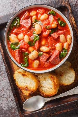 Herzhafter Bohnen-Eintopf mit Würstchen, Spinat und Gewürzen in Tomatensauce hautnah in einer Schüssel serviert mit geröstetem Brot auf dem Tisch. Vertikale Ansicht von oben
