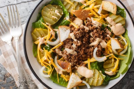 American Cheeseburger Salad with romaine lettuce, ground beef, pickles, cheddar cheese, onions and dressed with a special burger sauce closeup on the bowl on the table. Horizontal top view from abov