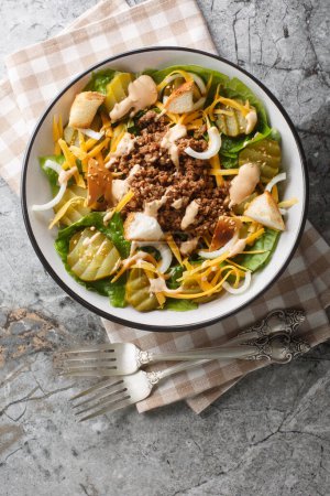 Cheeseburger salad with romaine lettuce, ground beef, pickles, cheddar cheese and croutons close-up in a bowl on the table. Vertical top view from abov
