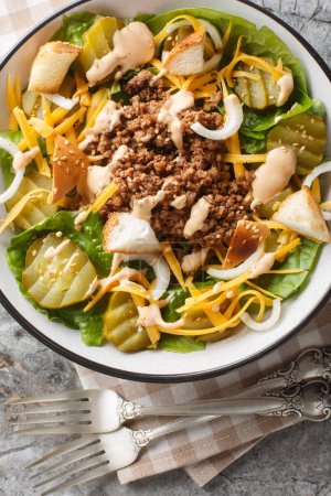 American salad with romaine lettuce, ground beef, pickles, cheddar cheese and croutons with sesame seeds close-up in a bowl on the table. Vertical top view from abov