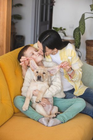 Téléchargez les photos : Un garçon en pull tricoté pose sur un canapé jaune avec sa mère et son chiot du Labrador - en image libre de droit