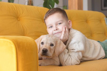 Téléchargez les photos : A boy in a knitted sweater poses on a yellow sofa with his Labrador puppy - en image libre de droit