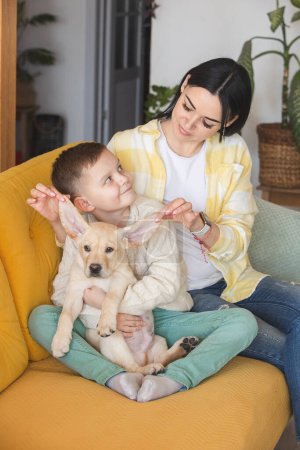 Foto de A boy in a knitted sweater poses on a yellow sofa with his mom and Labrador puppy - Imagen libre de derechos