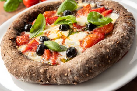 Téléchargez les photos : Délicieuse pizza noire à la pâte de céréales et de charbon de bois, garnie de mozzarella, sauce, olives noires et feuilles de basilic frais - en image libre de droit