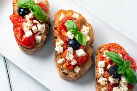 Photo for Delicious bruschette topped with mozzarella, olives, fresh tomatoes, basil, oregano and olive oil, typical italian snack, mediterranean diet - Royalty Free Image