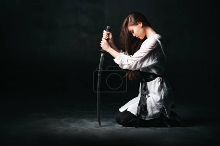 Téléchargez les photos : Ninja samurai woman kneeling and sitting on the floor, holding katana sword. The concept of loyalty and fidelity. Background with copy space. - en image libre de droit