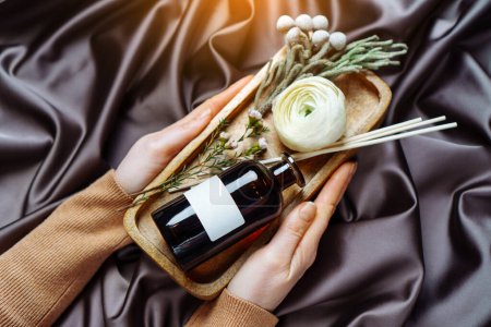 Female hands holding tray with scented home perfume in glass jar with rattan sticks. Luxury aroma oil with woody and flowers fragrance concept.