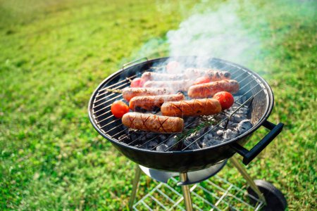 Photo for Grilled sausages grill. Roasting sausages bbq with tomatoes and rosemary in a round grill in a meadow. Summer picnic outdoors. - Royalty Free Image