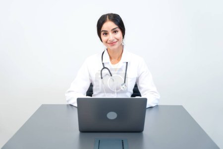Pretty indian woman GP doctor in uniform sitting at the laptop. Online medicine, remote consultation and health care.