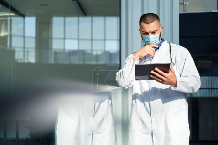 A serious and attentive male doctor wearing a white lab coat is using a tablet computer with focus and dedication in a hospital setting, showcasing professionalism and modern healthcare technology.
