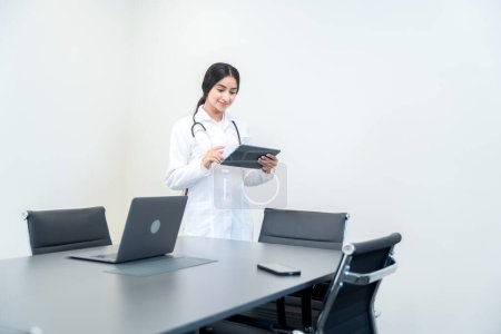 Doctora india joven vestida con una bata blanca de laboratorio y estetoscopio, parada junto a la mesa, consultando con un paciente en línea usando una tableta. 