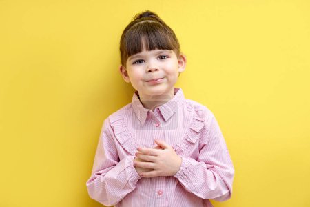 Téléchargez les photos : Girl Keep Hands On Chest, Thankful For Something, Feeling Gratitude. Sweet Child Of Caucasian Appearance Posing Isolated Over Yellow Background. Funny Girl Smiling - en image libre de droit