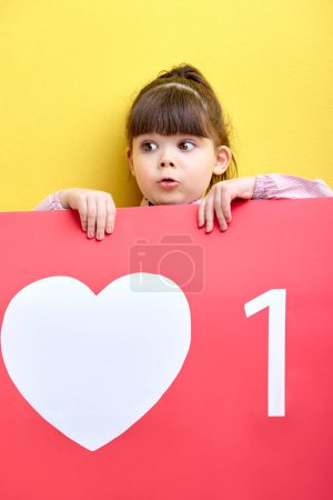 Foto de Funny kid girl holding big red sign like, posing, little blogger get likes, new followers on blog. isolated yellow background, surprised by raising followers in blog - Imagen libre de derechos