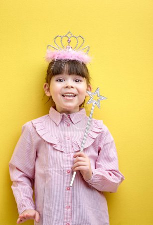 Foto de Happy smiling girl with crown on head, holding magic wand, looking at camera, yellow studio wall, copy space. little fairy posing, having caucasian appearance - Imagen libre de derechos